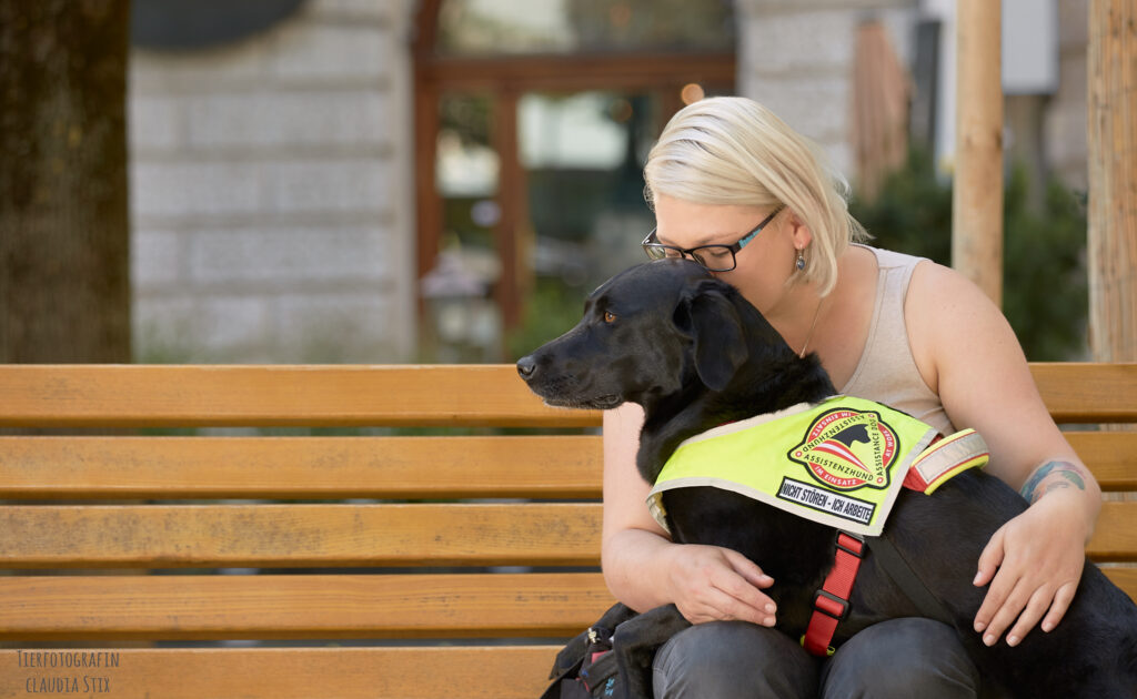 Eine junge Frau sitzt auf einer Parkbank und auf ihrem Schoß liegt mit dem Oberkörper ein schwarzer Labrador Retriever. Der Hund trägt eine gelbe Kenndecke, die ihn als Assistenzhund ausweist.